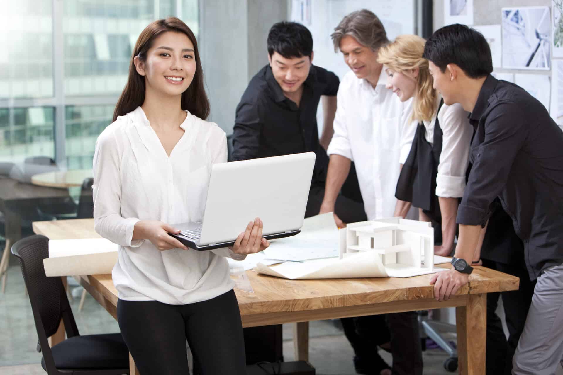 Chinese architect holding laptop in studio with her team in background